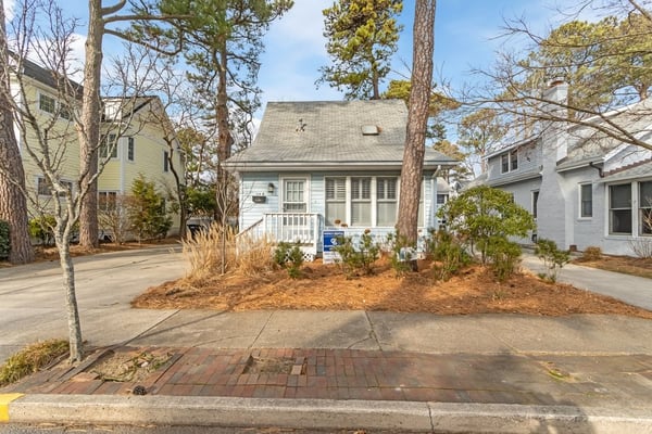 White cottage with gray roof and front porch, surrounded by tall pine trees and mulched landscaping, brick driveway visible.
