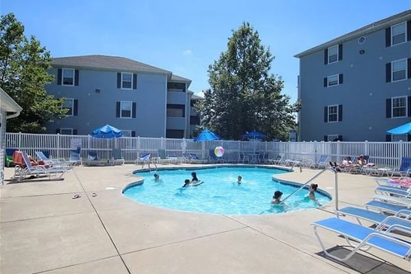 Outdoor swimming pool with several swimmers, surrounded by white fence, lounge chairs, and two-story residential buildings under clear blue sky