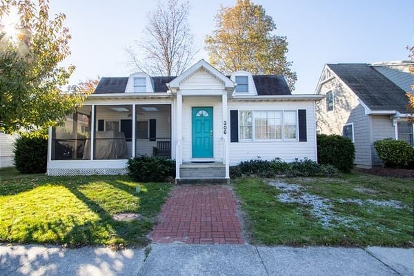 White cottage-style house with turquoise front door, screened porch, and brick walkway in autumn setting