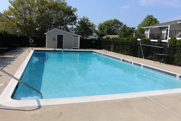Community swimming pool with clear blue water, concrete deck, metal railings, utility shed, and residential homes in background