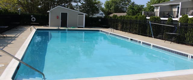 Community swimming pool with clear blue water, concrete deck, metal railings, utility shed, and residential houses in background surrounded by trees