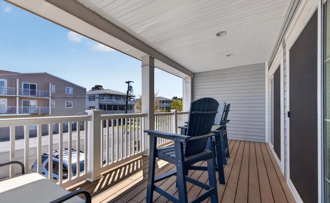 Covered deck with dark blue Adirondack chair, white railings, and view of neighboring beach houses and clear sky