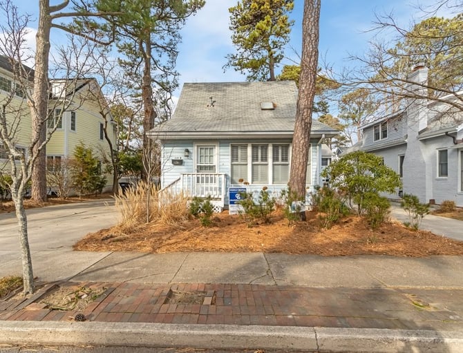 White cottage with gray roof and front porch surrounded by tall pine trees and mulched landscaping, brick driveway visible.